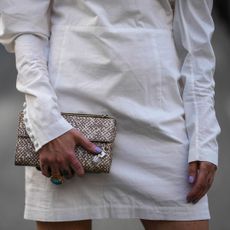 A crop of a woman wearing a white long-sleeved dress holding a snakeskin clutch bag and lavender nails