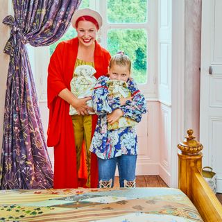 Angel and daughter Dorothy from Escape to The Chateau standing in bedroom in front of full length heavy floral purple curtains, both hugging a cosy patterned hot water bottle