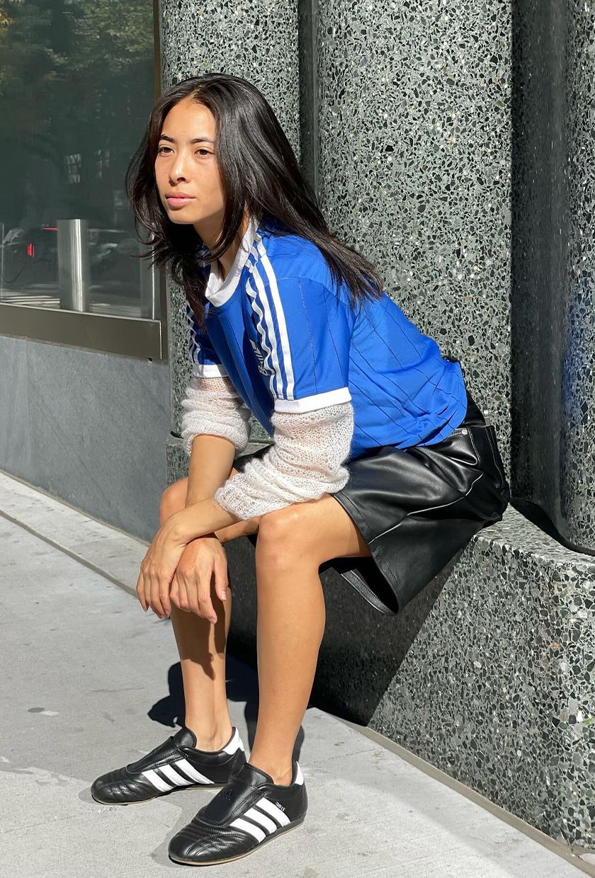 A woman wearing a blue short-sleeve sports jersey over a white long-sleeve gauzy knit shirt with leather Bermuda shorts and Adidas shoes.