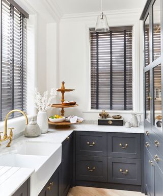 Modern white and ark blue kitchen, dark brown Venetian blinds, hanging glass pendant, countertops decorated with kitchenware and accessories