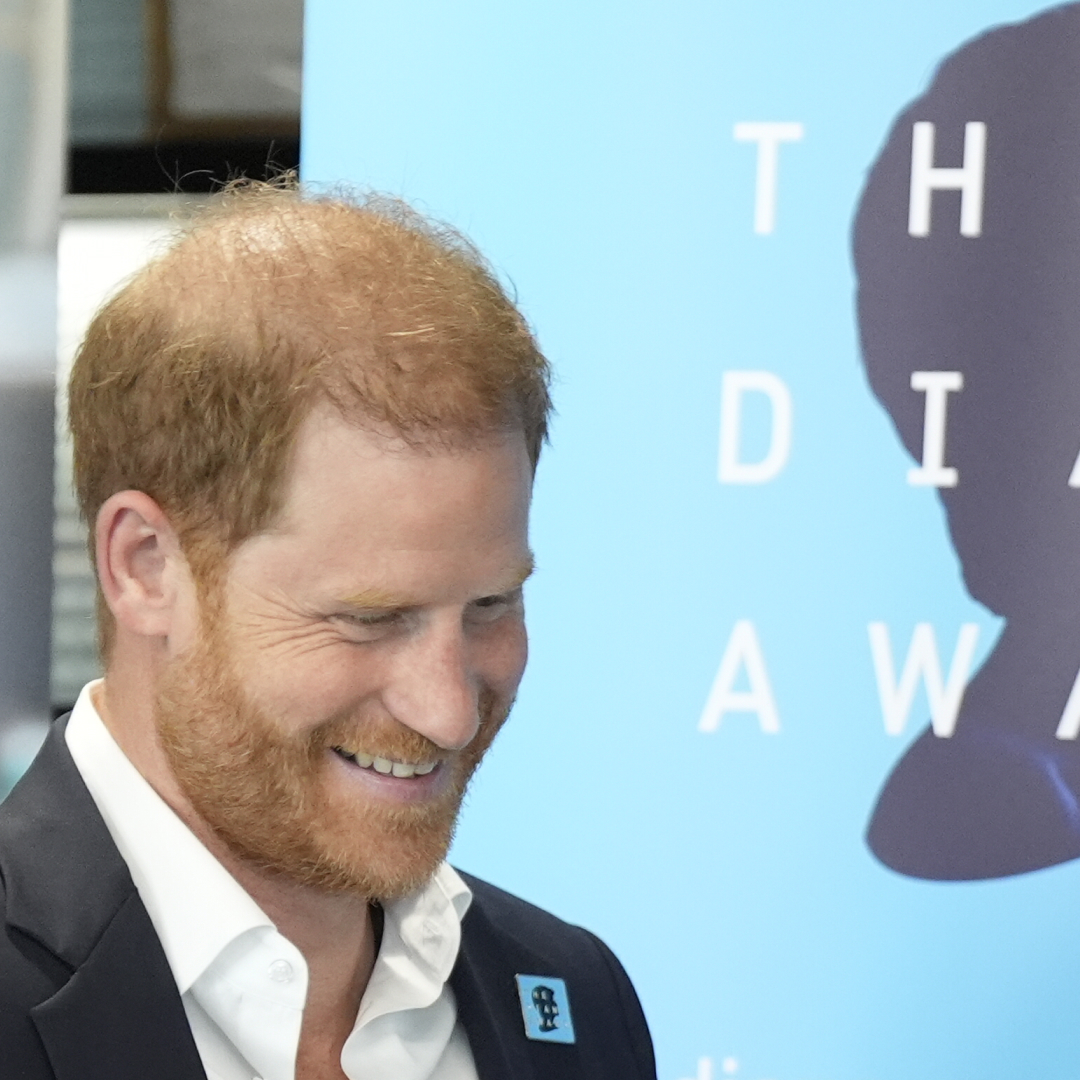 Prince Harry, in a suit and white shirt, smiling in front of a blue Diana Award backdrop