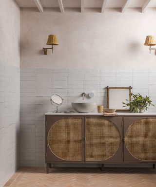 Rustic, natural bathroom with white wall tiles, terracotta floor tiles, cabinet with rattan doors, decorated with plants, wooden accessories, wall lamps with wicker woven shades.