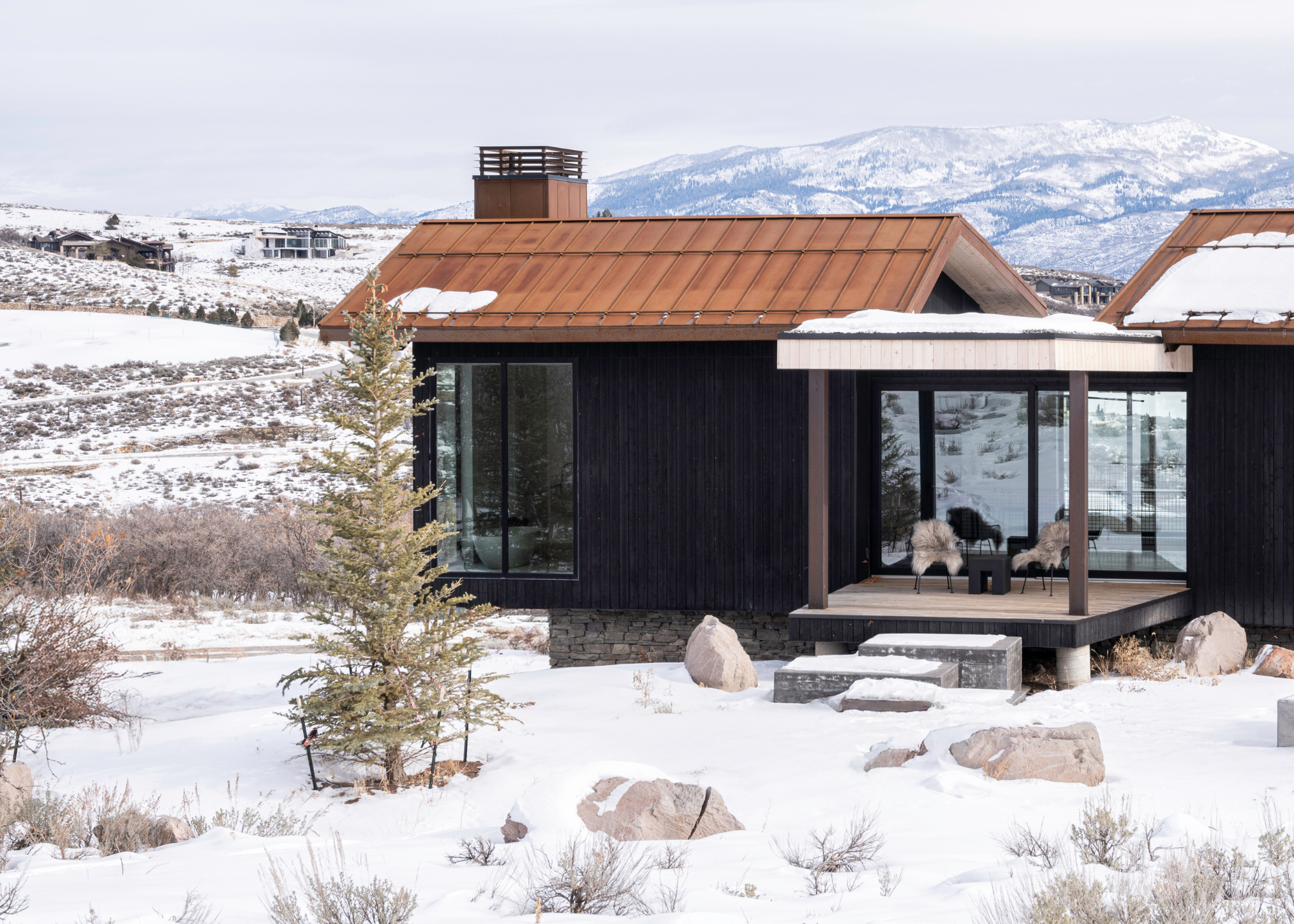 snowy cabin house exterior shot with a view of a decking area that also has seating and a table