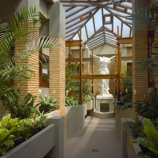Inside the Darwin D. Martin House conservatory, part of a wider complex built between 1903 and 1905. A indoor passage with planters on either side of it, a white statue at the end and wood and glass roof above it.