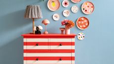 Image of a dresser painted in red and white stripes with two lamps and three vases on top of it. The back wall is a light blue with colorful plates hung on it.