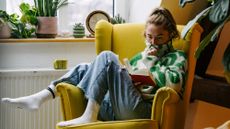 A woman sitting on a yellow armchair surrounded by plants in her living room and reading a book