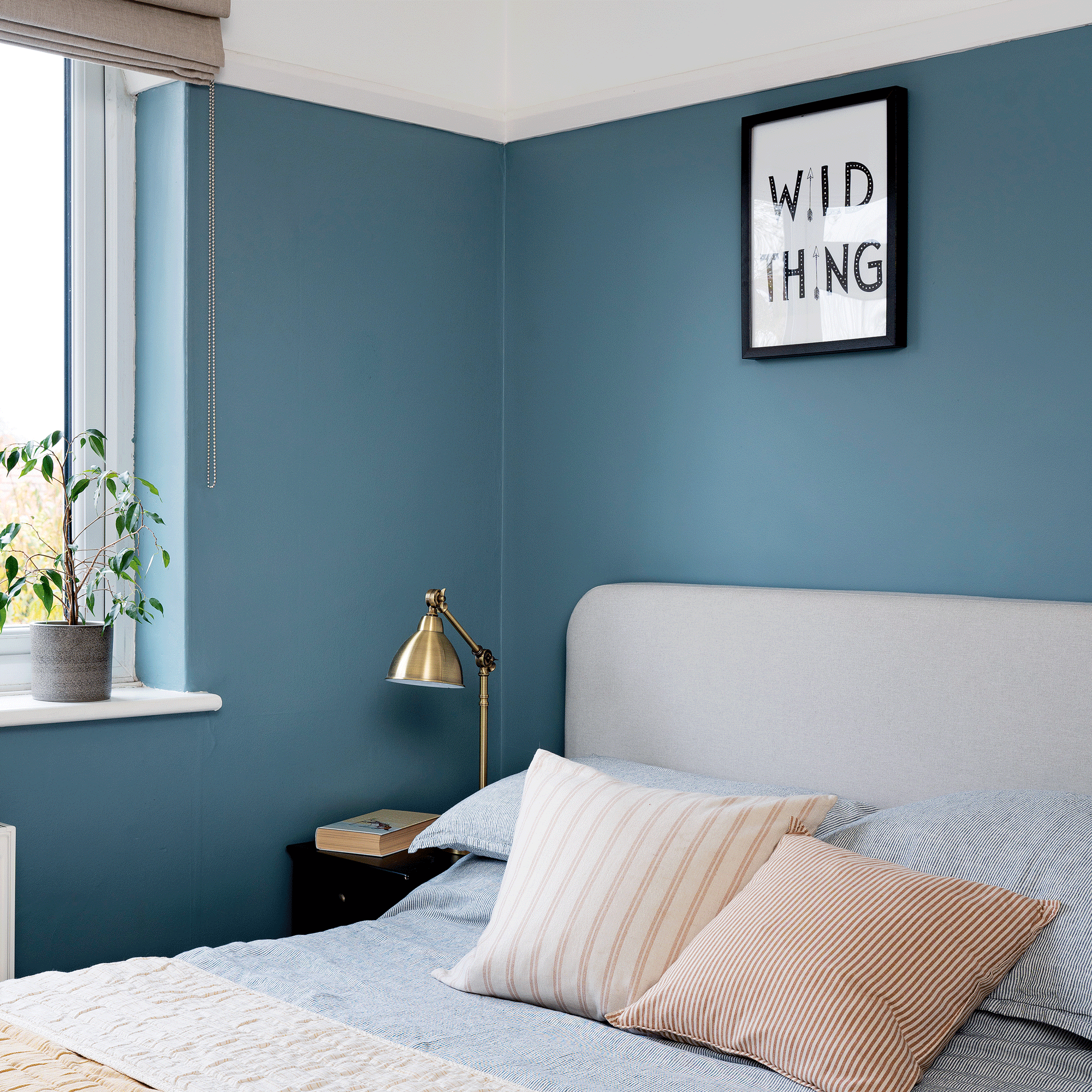 a bedroom with blue painted walls framed artwork striped cushions and a brass bedside lamp