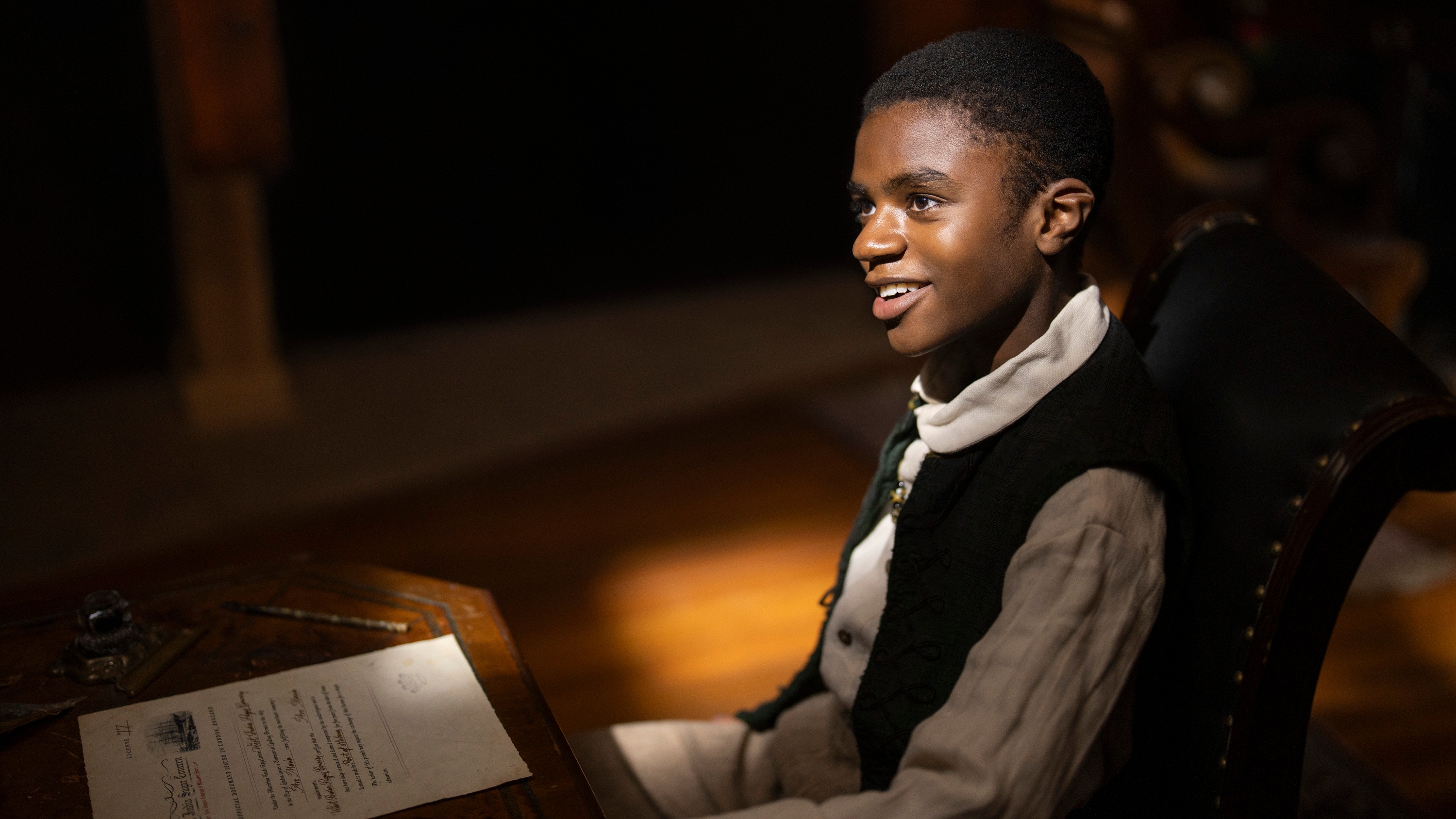 Eddie Karanja as Washington Black aged 11. He's seen here in period dress, in a dark room, smiling as the sunlight hits his face, and before him is a piece of paper.