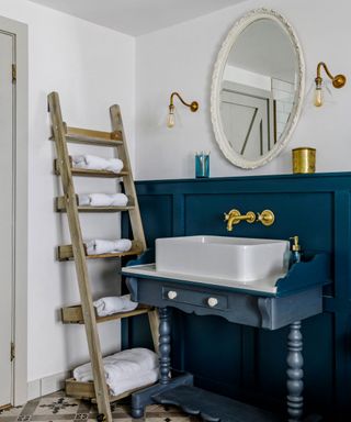 Traditional, country style bathroom with tiled flooring, blue painted paneling and matching, wooden sink unit, rounded mirror with cream frame, two metallic wall lights either side, wooden ladder acting as shelving unit, towels stored on each section