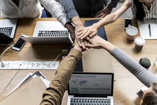 A team of colleagues joining hands over a desk scattered with laptops and smartphones