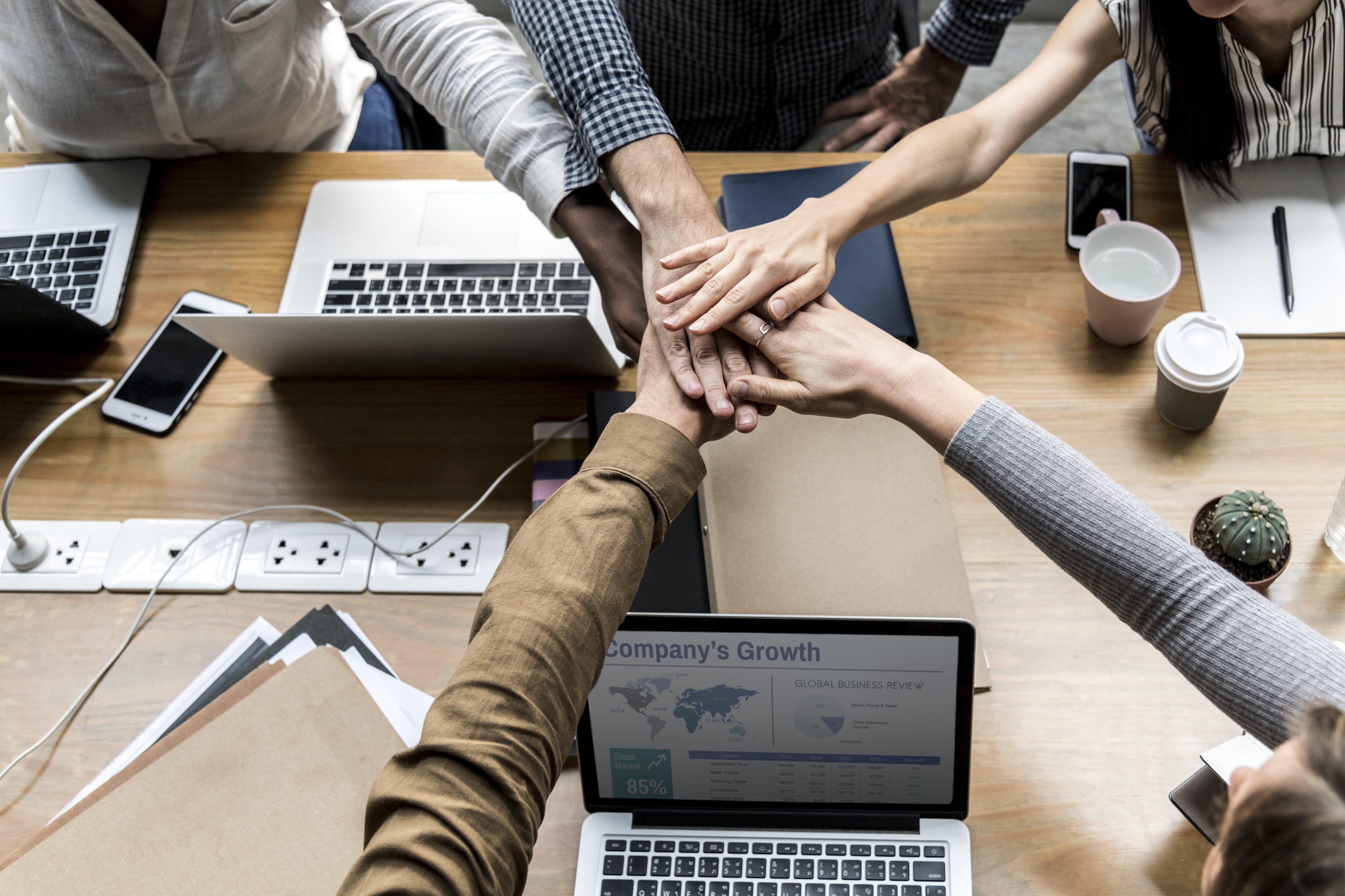 A team of colleagues joining hands over a desk scattered with laptops and smartphones