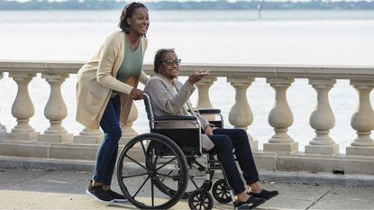 A daughter pushes her mom in a wheelchair while walking across a bridge.