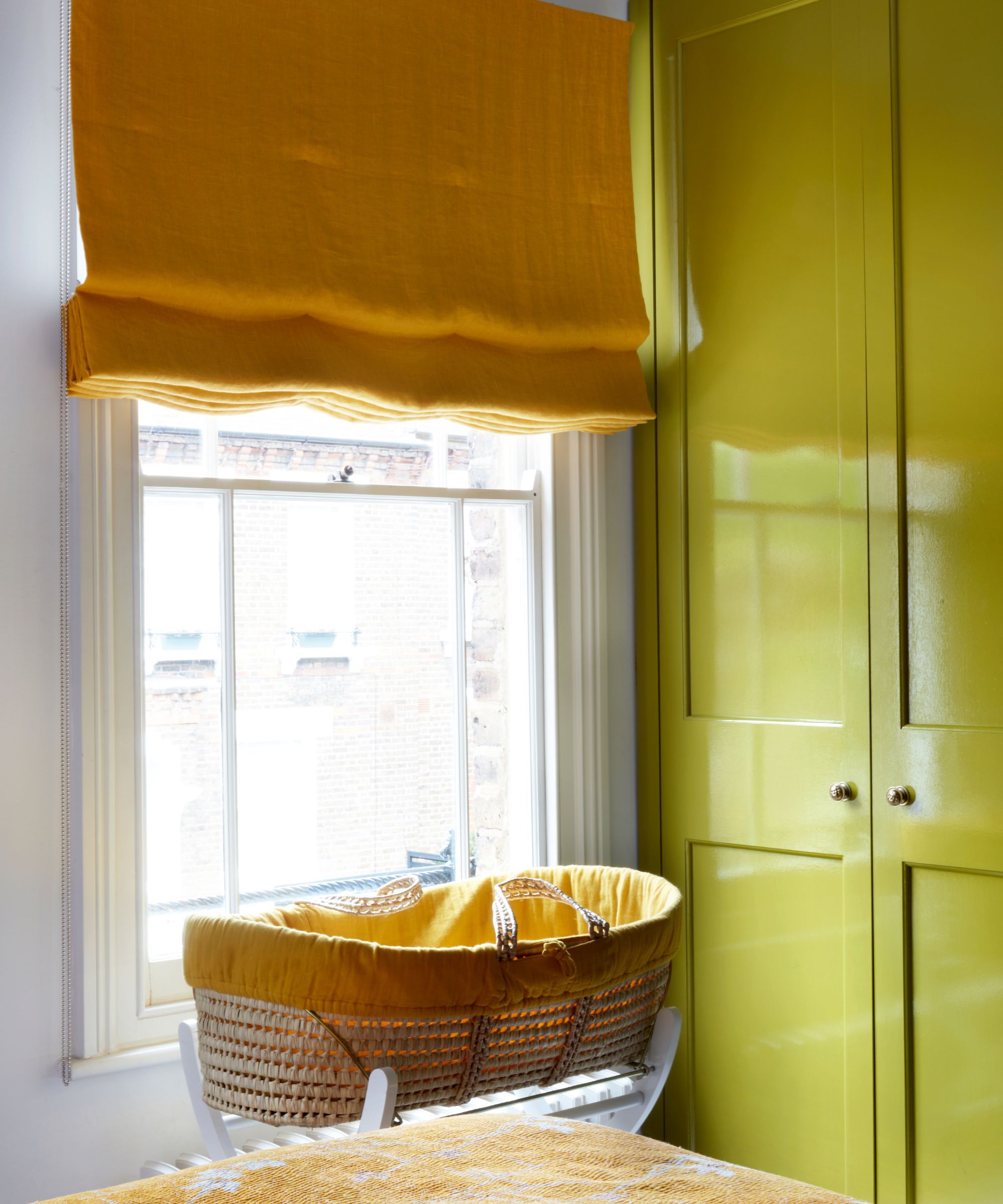 Bedroom with a citron green lacquer painted wardrobe, a large window with a yellow roman blind, a basket with a yellow lining underneath the window and a bed with a yellow wool throw.