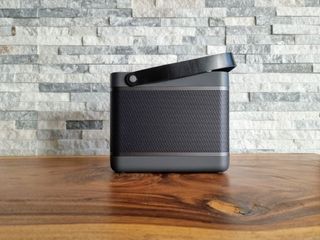 En grå Bluetooth-högtalare av typen Bang & Olufsen Beoli 20 med ett svart handtag står på ett träfärgat bord mot en grå tegelvägg.