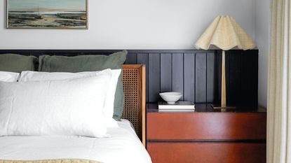 White bedroom with rattan bed with white and green bedding, black wall panelling, wood chest of drawers, and scalloped lamp with a landscape artwork above
