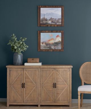 dark blue wall with wooden cabinet with vase of foliage, two framed artworks and rattan accent chair