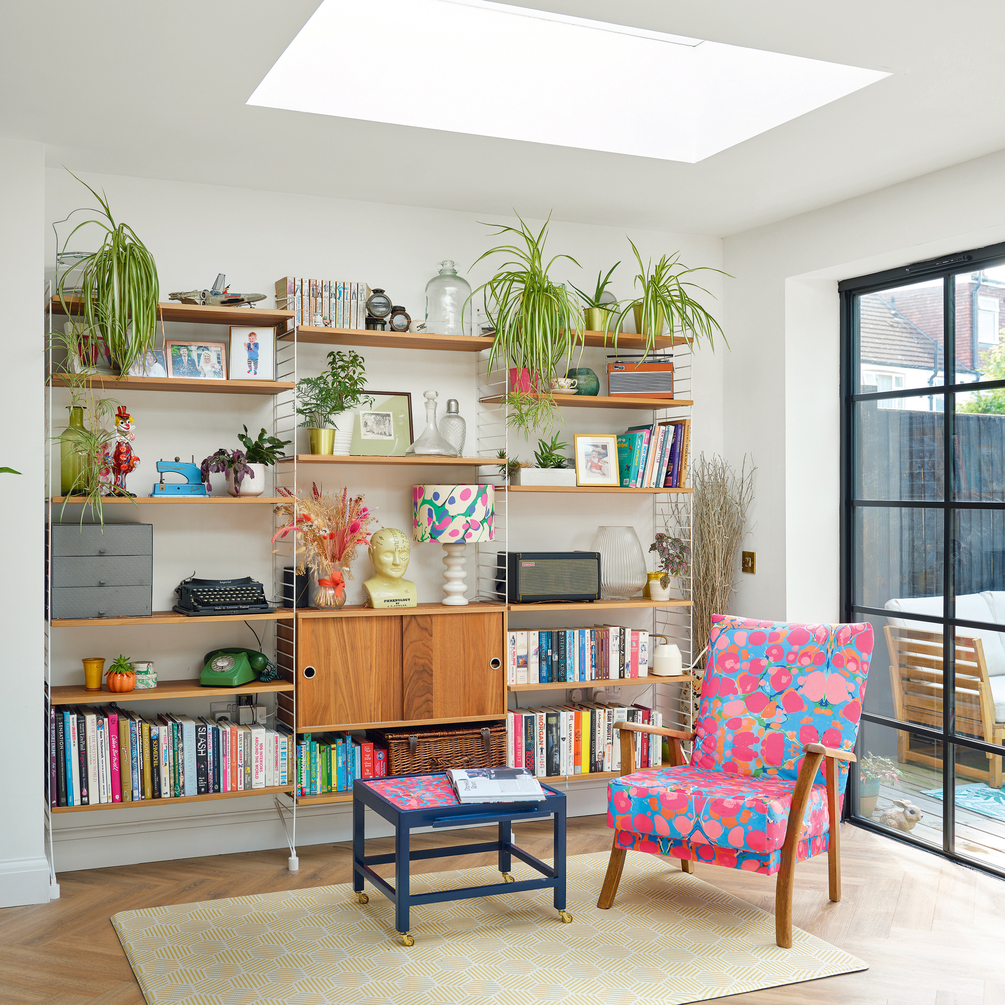 a mid-century wall-hung shelving unit filled with books, plants, and accessories with a brightly coloured armchair beside it