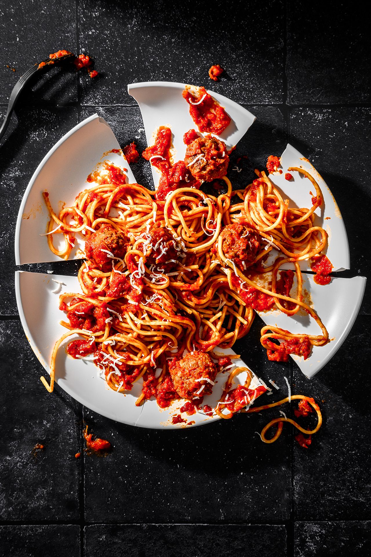 A broken plate with tangled spaghetti and meatballs in marinara sauce, scattered sauce splatters, on a dark surface