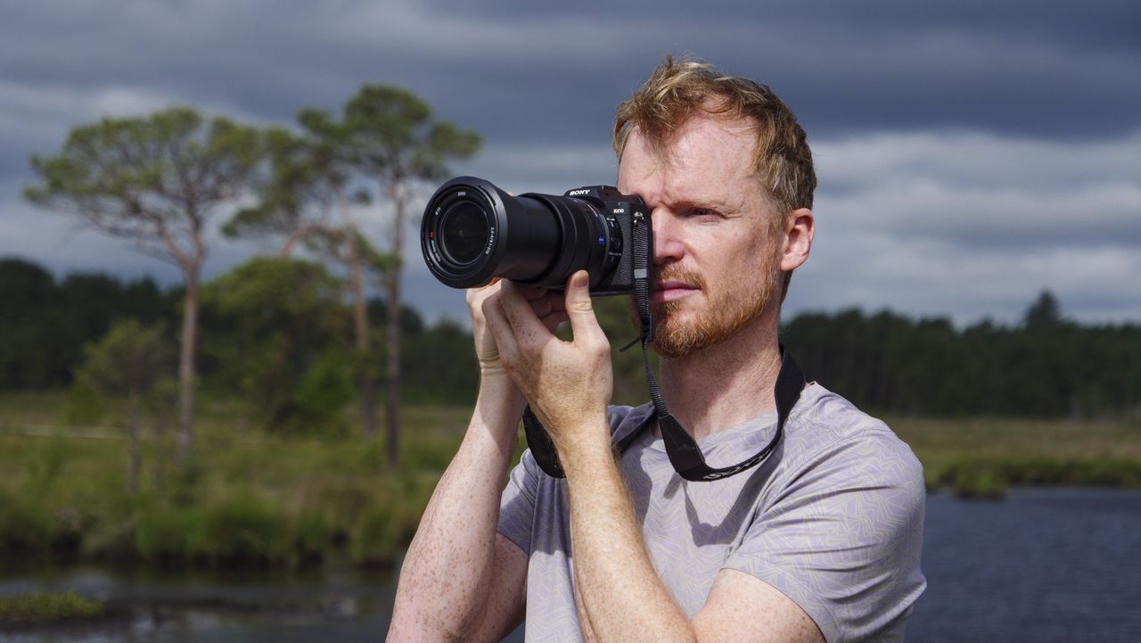 Photographer and tech journalist Tim Coleman holding the Sony RX10 V bridge camera up to his eye in a nature reserve, with dramatic clouds behind him