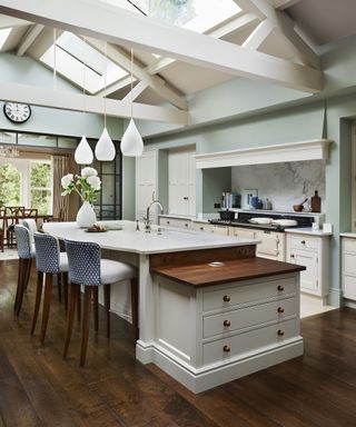 A curved island, upholstered bar stools and three pendant lights in a large cream kitchen with high a ceiling and wooden beams.