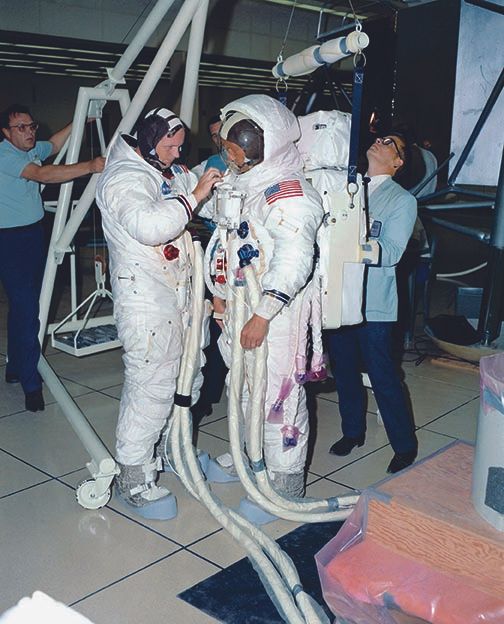 Armstrong and Aldrin participate in an Extravehicular Mobility Unit donning procedure verification exercise in the Manned Spacecraft Operations Building at Kennedy Space Center on June 25. The procedure allowed the crewmembers to have a final look at the flight hardware and gave them an opportunity to resolve any issues. Armstrong is connecting the Oxygen Purge System on/off actuator to the Remote Control Unit (RCU). On the left is Joe Johnson with the GE Support Group, with Jim Howe of Hamilton Standard on the right.