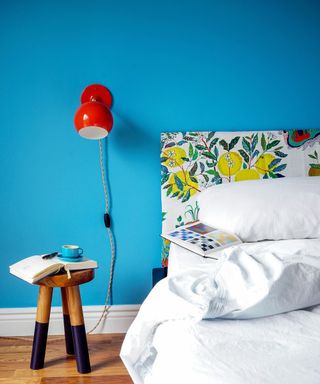 bright blue wall in bedroom with white bedding, floral headboard and red wall light with wooden stool as nightstand