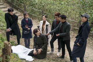 a group of people (l-r, Sehun, Kim Se-jeong, Park Min-young, Kim Jong-min, Ahn Jae-wook, Yoo Jae-suk) stand over a man (Lee Kwang-soo, center) as he kneels over a corpse covered by a white sheet, in the netflix show busted