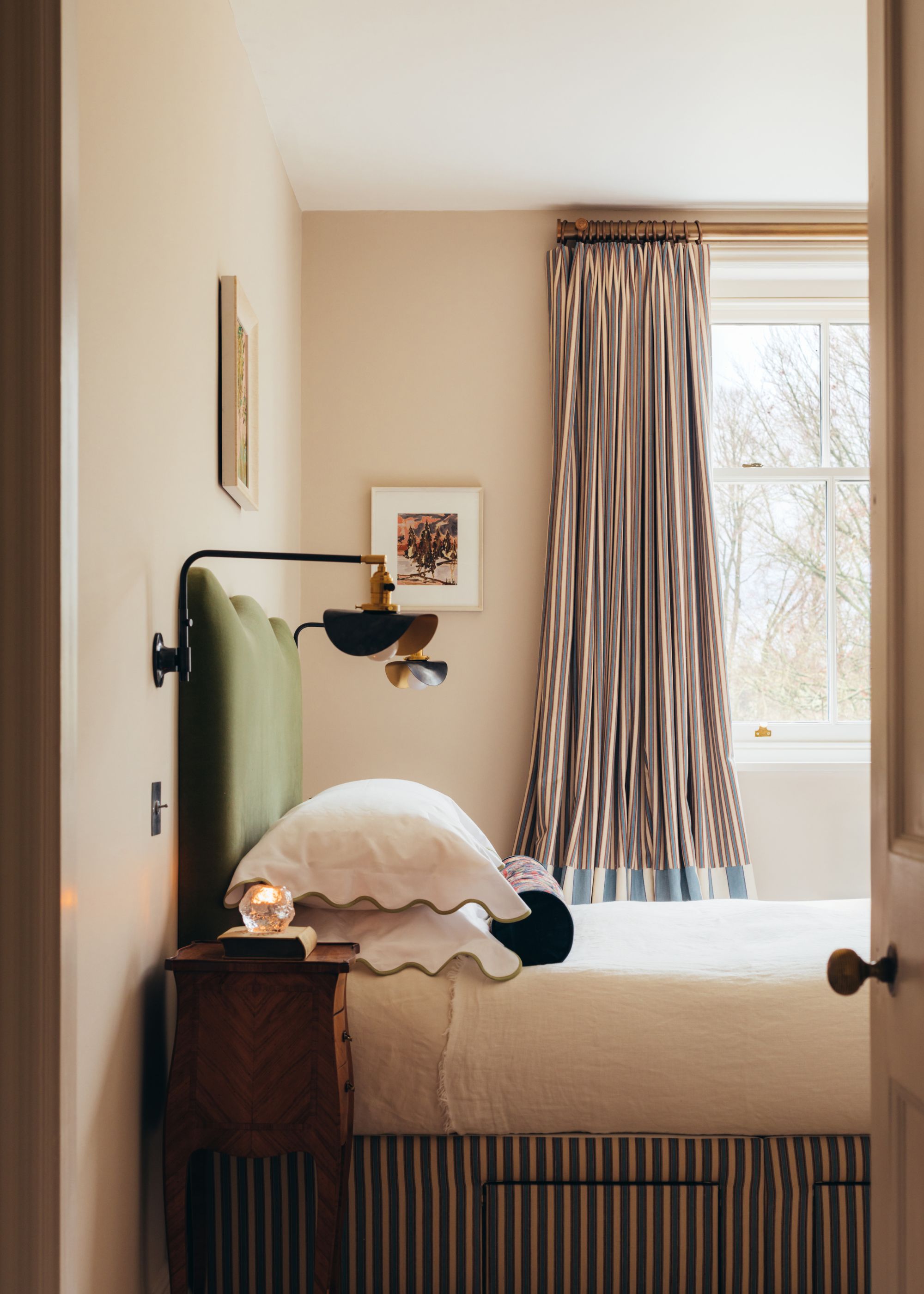 A bedroom with soft beige walls, striped curtains, white bedding with scalloped pillows, and a green headboard with two wall lights.