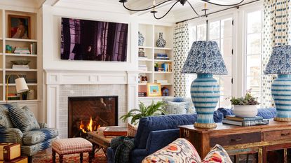 Living room with white walls, built-in bookcases with books and decor, fireplace with TV mounted above, blue sofa, red Persian rug, and console behind sofa with lamps and books
