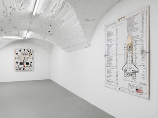 View of two pieces of wall art. One features an image of a rocket, the American flag and small black text and the other features mixed media and the wording 'TRAINING' in black. The pieces are displayed in a room with grey flooring, white walls and a white arched ceiling