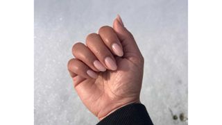 A close-up of Digital Beauty Editor, Aleesha Badkar's hand with sheer pink almond nails, pictured in front of a snowy backdrop