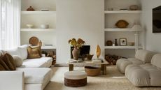Image of an off-white living room with a white sectional and two boucle accent chairs. The back wall has open shelving on either side of a fireplace. There is a marble coffee table in the middle.