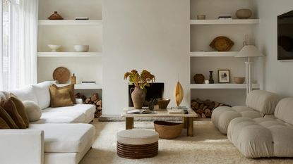 Image of an off-white living room with a white sectional and two boucle accent chairs. The back wall has open shelving on either side of a fireplace. There is a marble coffee table in the middle.