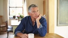 A middle-aged man sits at the table with a cup of coffee, chin in hand. He looks sad or thoughtful.