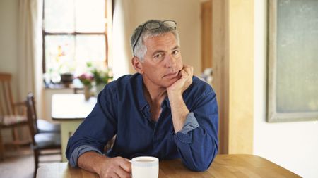 A middle-aged man sits at the table with a cup of coffee, chin in hand. He looks sad or thoughtful.