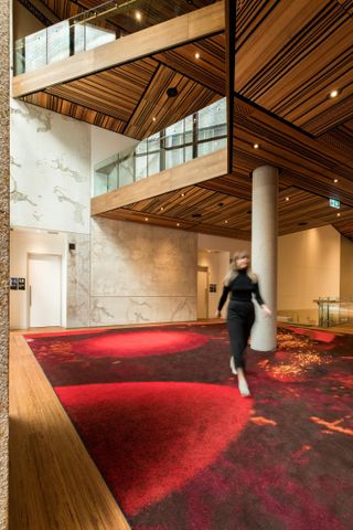 Foyet interior, Red patterned carpet, Lady dressed in black walking creating a blurred image, Marble stone wall and stone pillars leading up to wooden and glass gallery with spotlights