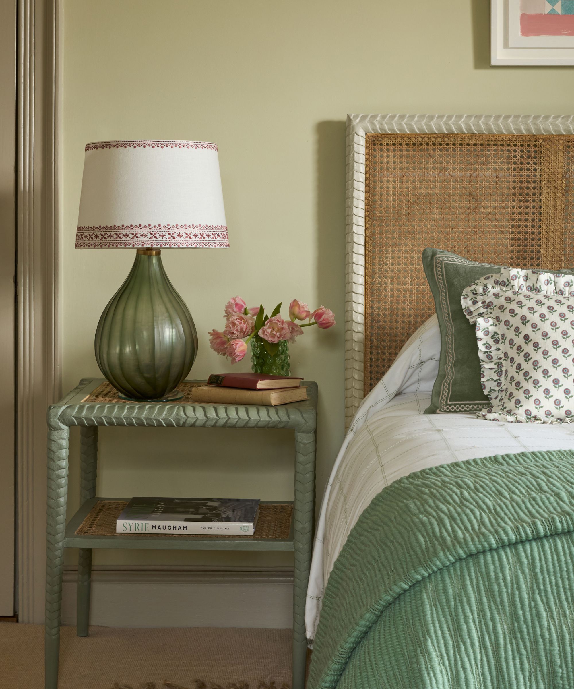 A bedroom painted sage green with a woven bed-frame, green and white bedding, a sage green nightstand with a green glass lamp, and a small vase of flowers.