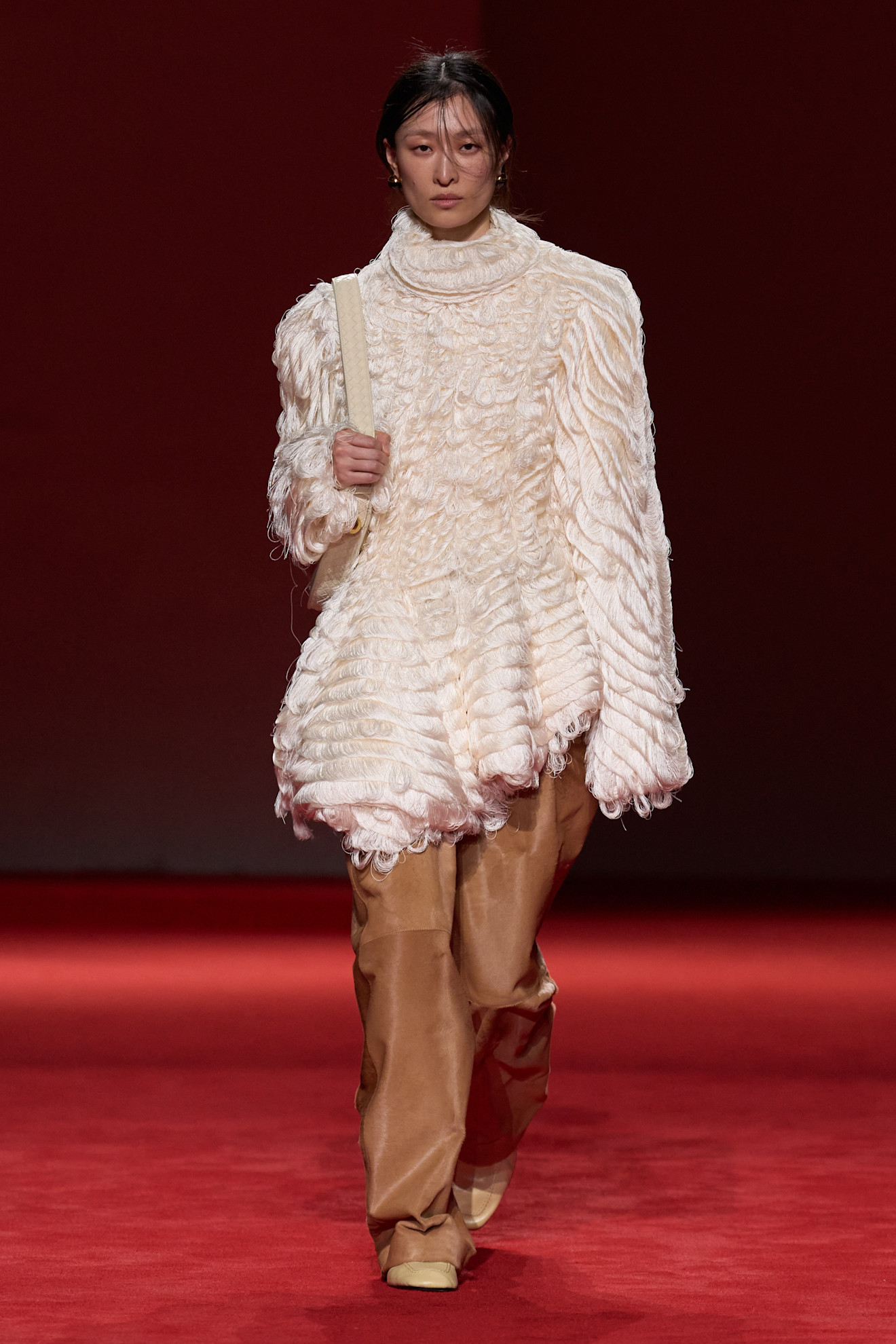 A model walking in the F/W 26 Bottega Veneta show during Milan Fashion Week wearing a textured turtleneck tunic with tan pony hair pants.