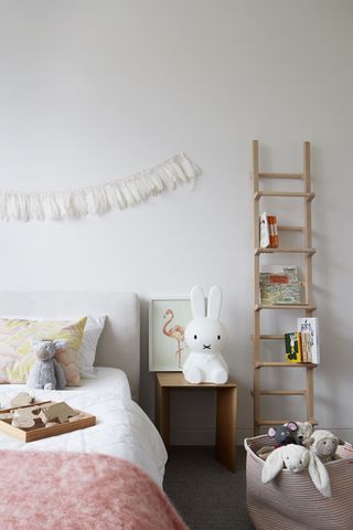 girl's bedroom with white walls, ladder storage, wooden modern bedside, feather bunting, pink and yellow bedspread/pillow, toys, basket for toys