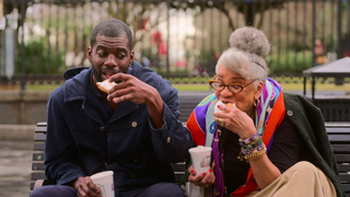 Stephen Satterfield and Jessica B. Harris, taking bites of beignets while sitting on a bench outside, in 'High on the Hog: How African American Cuisine Transformed America'