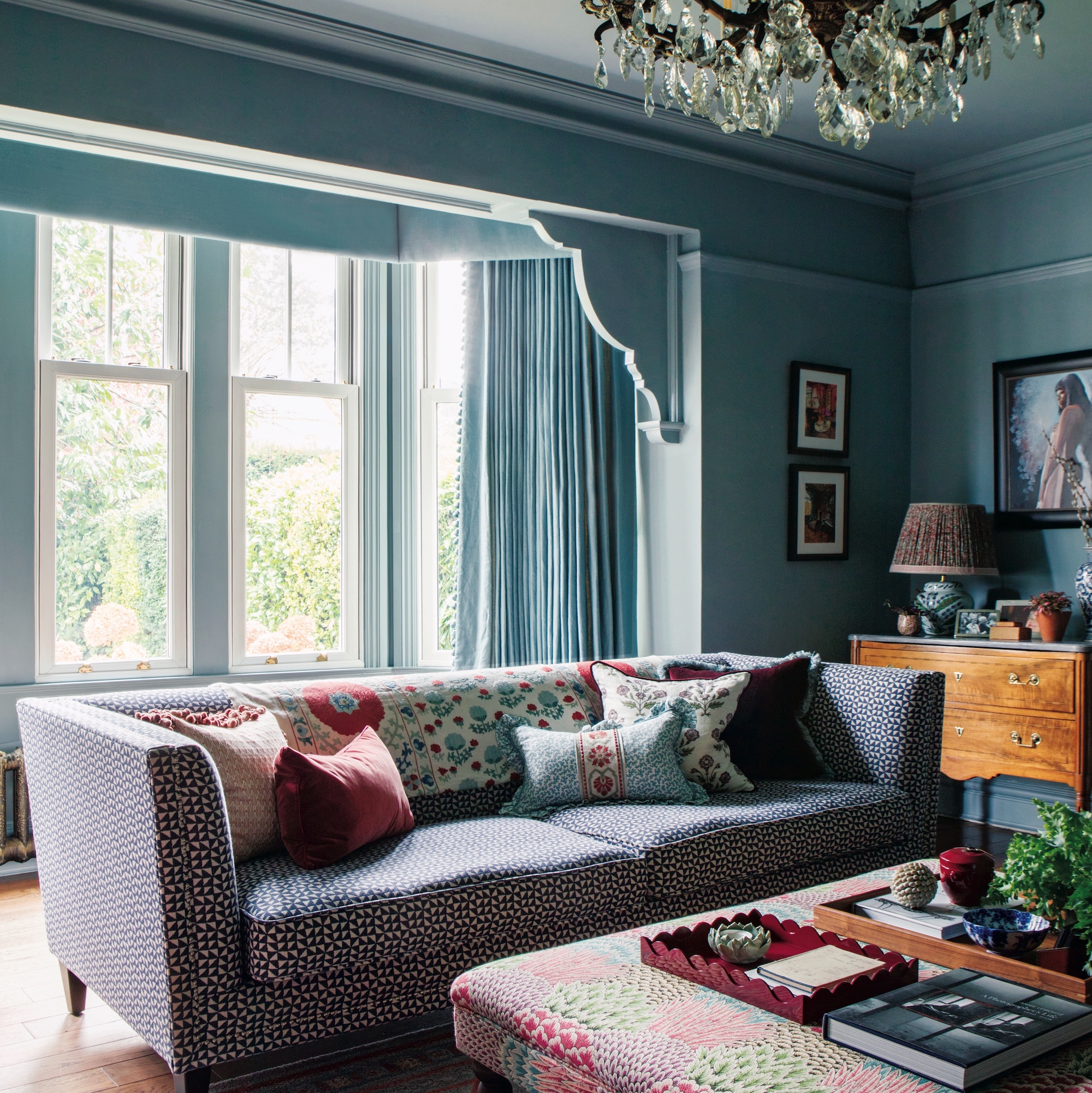 sitting room with blue walls and large crystal chandelier above a patterned sofa and ottoman with trays on the top