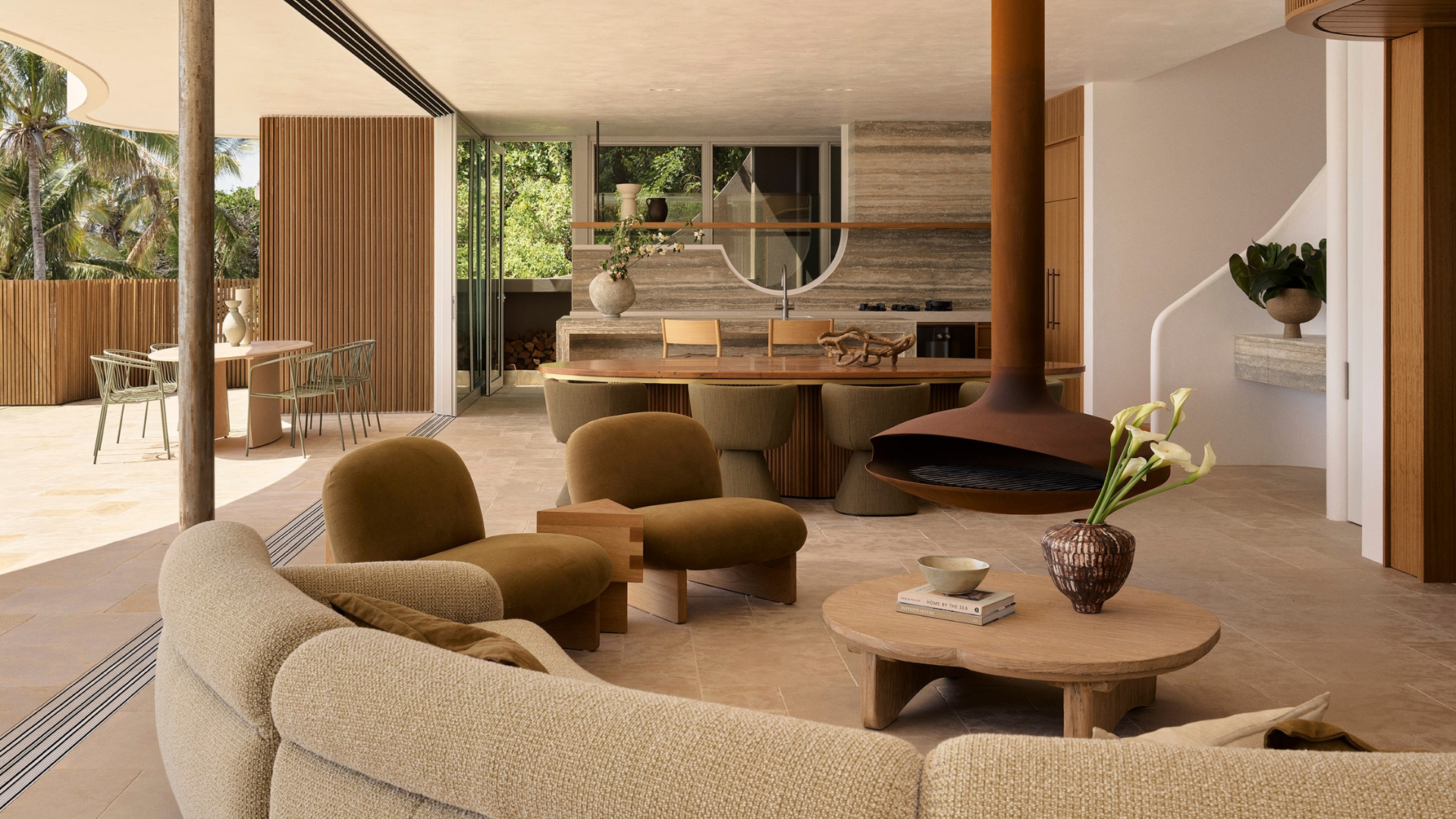 An earthy living room with a curved sectional sofa, brown suede chairs, a wood coffee table, a vase of tulips, a brown hanging fireplace, a stone kitchen, and the balcony doors drawn open