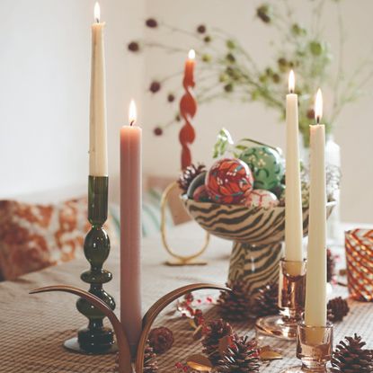 A dining table covered with a brown gingham tablecloth and decorated with various dinner candles and a bowl of Christmas baubles