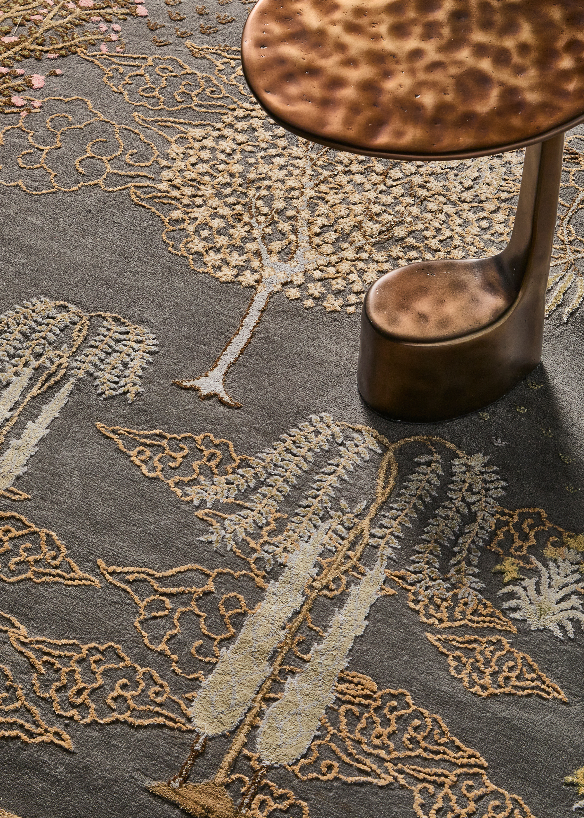 an abstract side table in punched brass with a thick base at the bottom that goes up thin on one side on top of an ornate japanese style rug