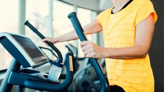 Woman holding onto handlebars of elliptical trainer at gym wearing yellow t-shirt