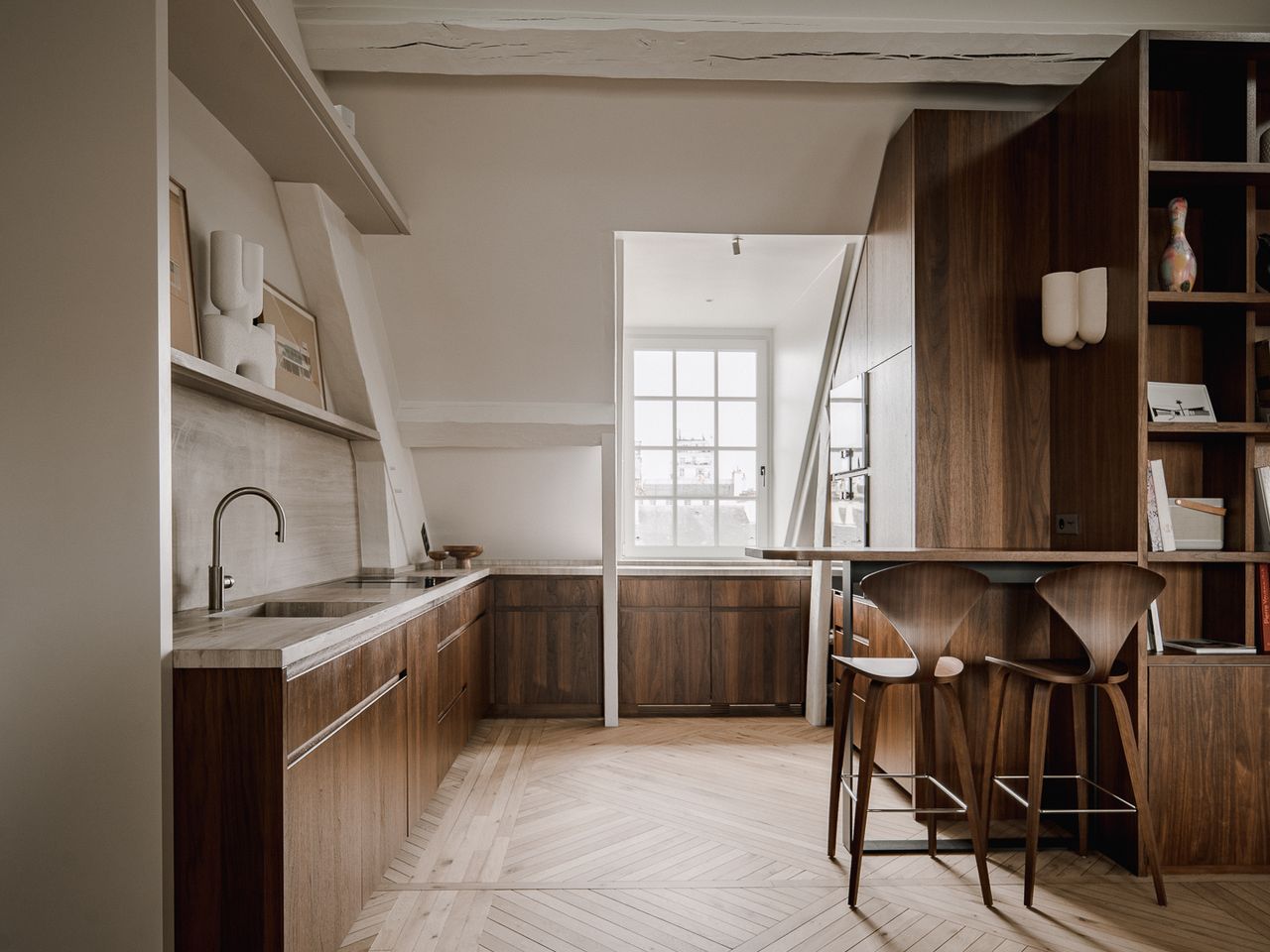 A refined attic apartment interior characterized by a warm, modernist aesthetic. The space is organized as an open-plan living area under a pitched roof, with exposed white-painted beams and triangular dormer windows that bring in soft natural light.