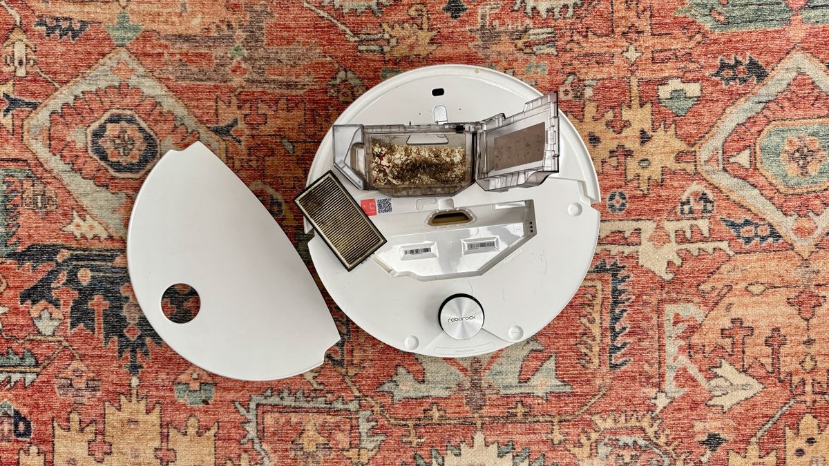 Roborock Qrevo Curv 2 Pro on a patterned carpet rug, with its onboard bin open to reveal oats and crumbs that it has just sucked up
