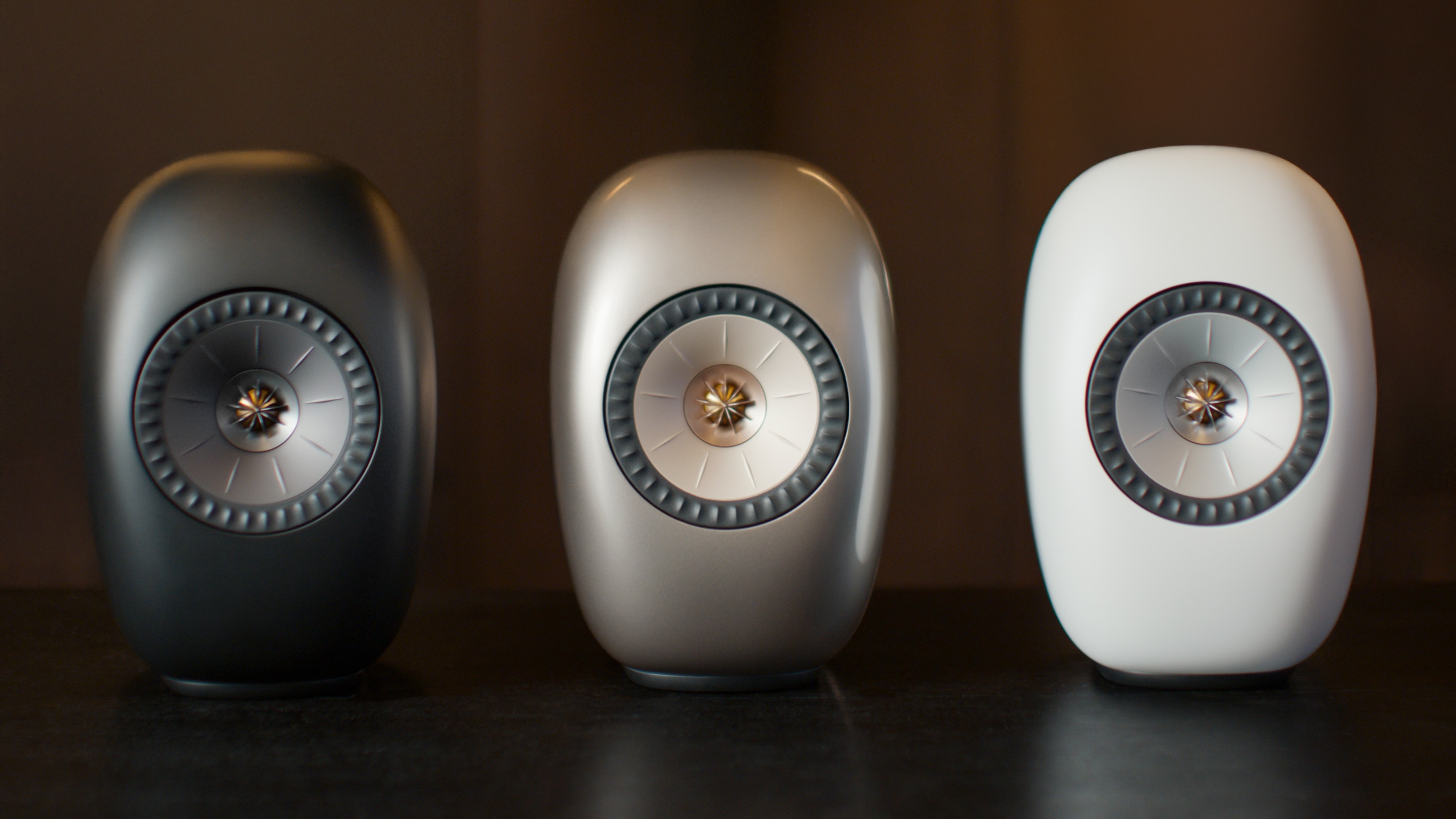 Three KEF LS Luxe speakers &ndash; in black, grey and white &ndash; in a row on a dark surface.
