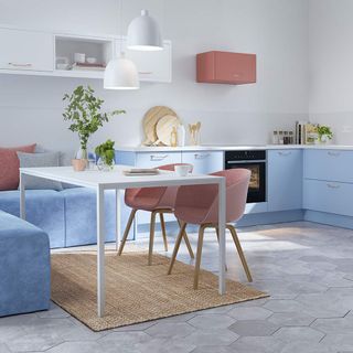 a kitchen with white walls and light grey hexagonal floor tiles, blue kitchen cupboards, a white dining table with pink chairs on one side and an L shaped blue sofa on the other