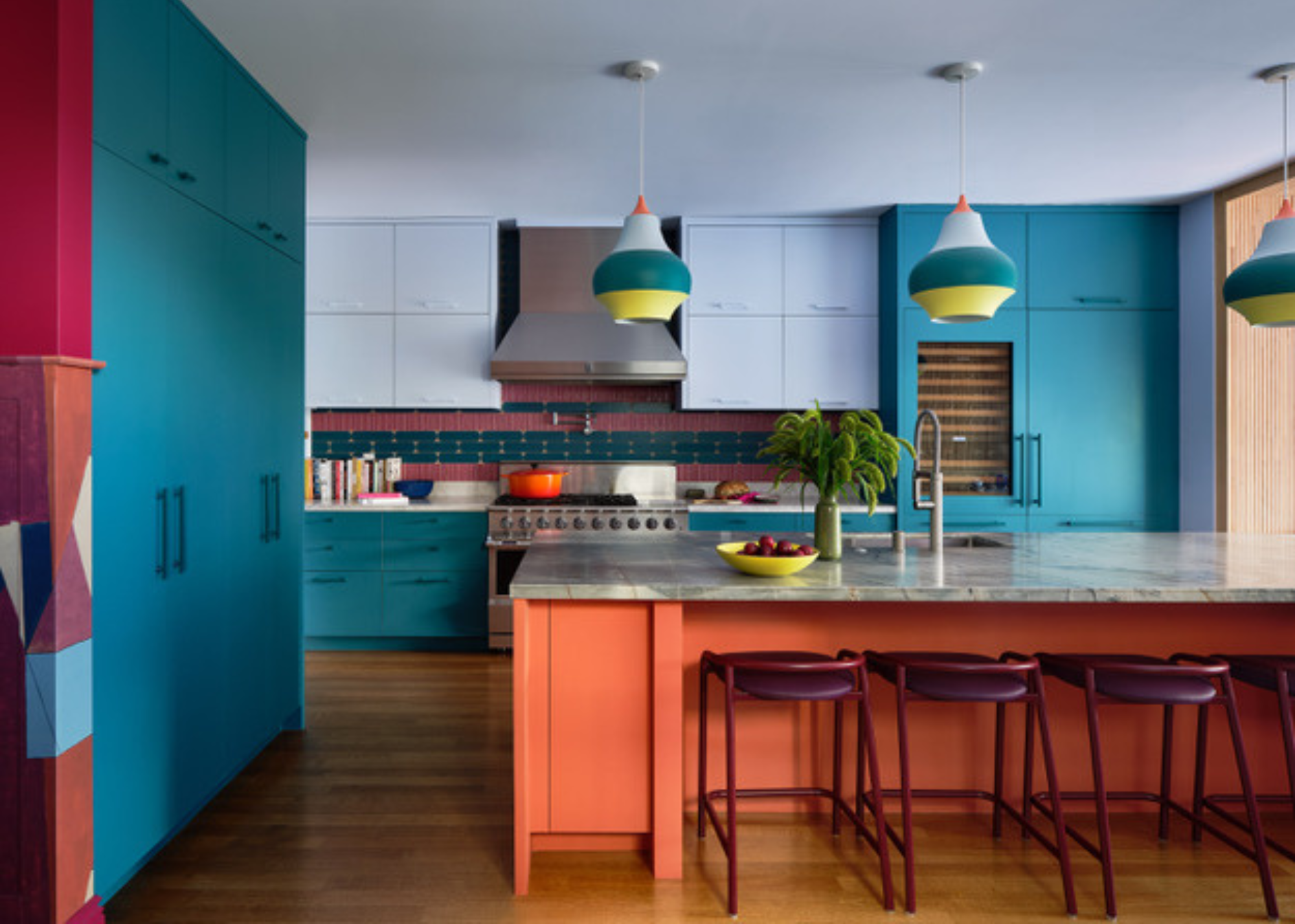 A playful modern teal kitchen with a coral island and grey countertop with burgundy bar stools tucked underneath. The island pendant lighting is also playful and ties in the color palette.